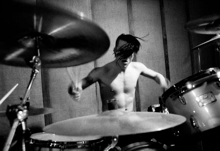 A shirtless adult male is playing a drum set with cymbals in a monochrome indoor music studio, playing the drums with drumsticks.