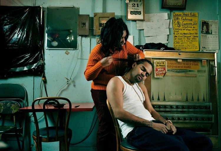 Woman braids another man's hair inside a room featuring chairs, tables, and wall decorations, illuminated by ambient lighting.