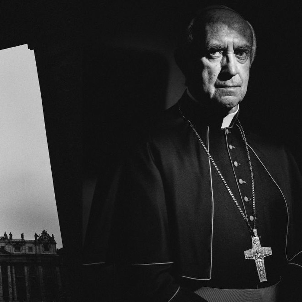 Portrait of an adult male in clerical collar and cross necklace, with St. Peter's Basilica visible in the background.