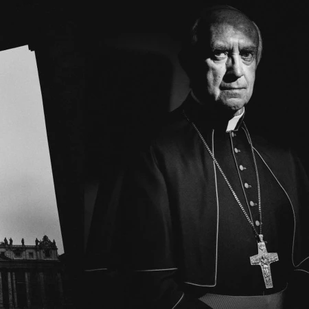 Portrait of an adult male in clerical collar and cross necklace, with St. Peter's Basilica visible in the background.