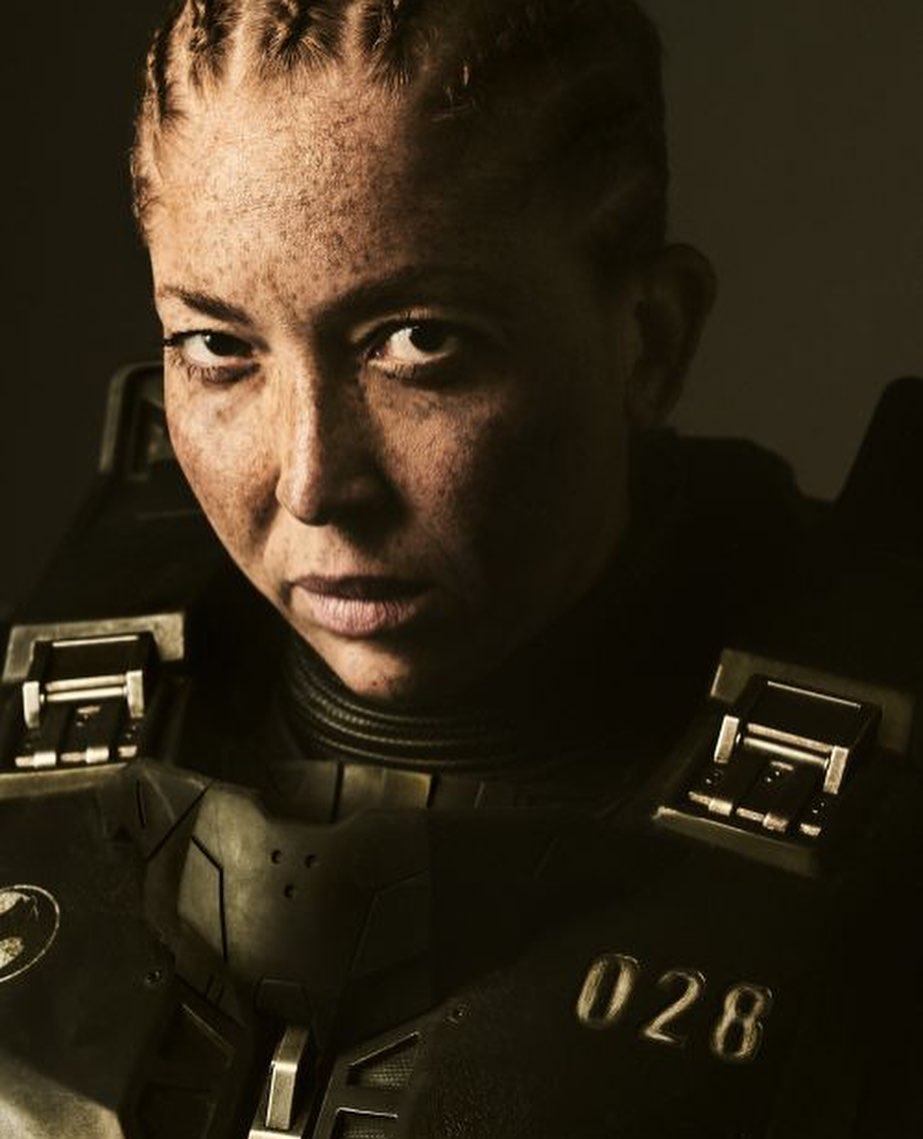 Close-up portrait of a female soldier with braided hair, wearing olive green military armor and stern expression, against a dark background.