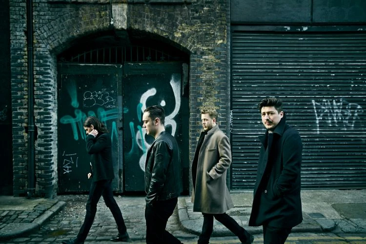 Four men in coats walk along a city street, posing in front of a weathered brick wall with graffiti, captured in a medium shot.