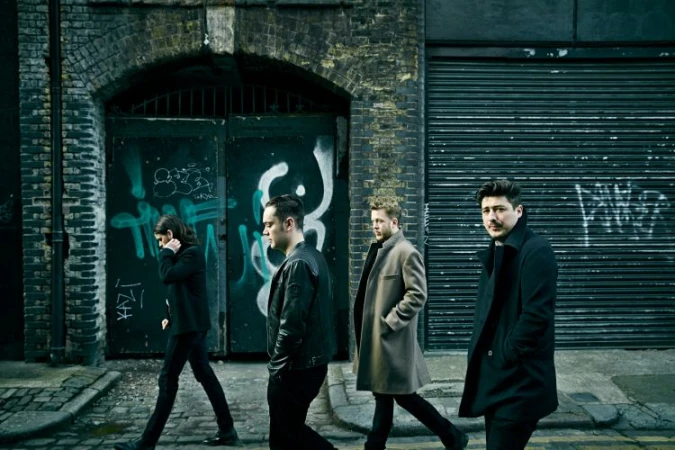 Four men in coats walk along a city street, posing in front of a weathered brick wall with graffiti, captured in a medium shot.