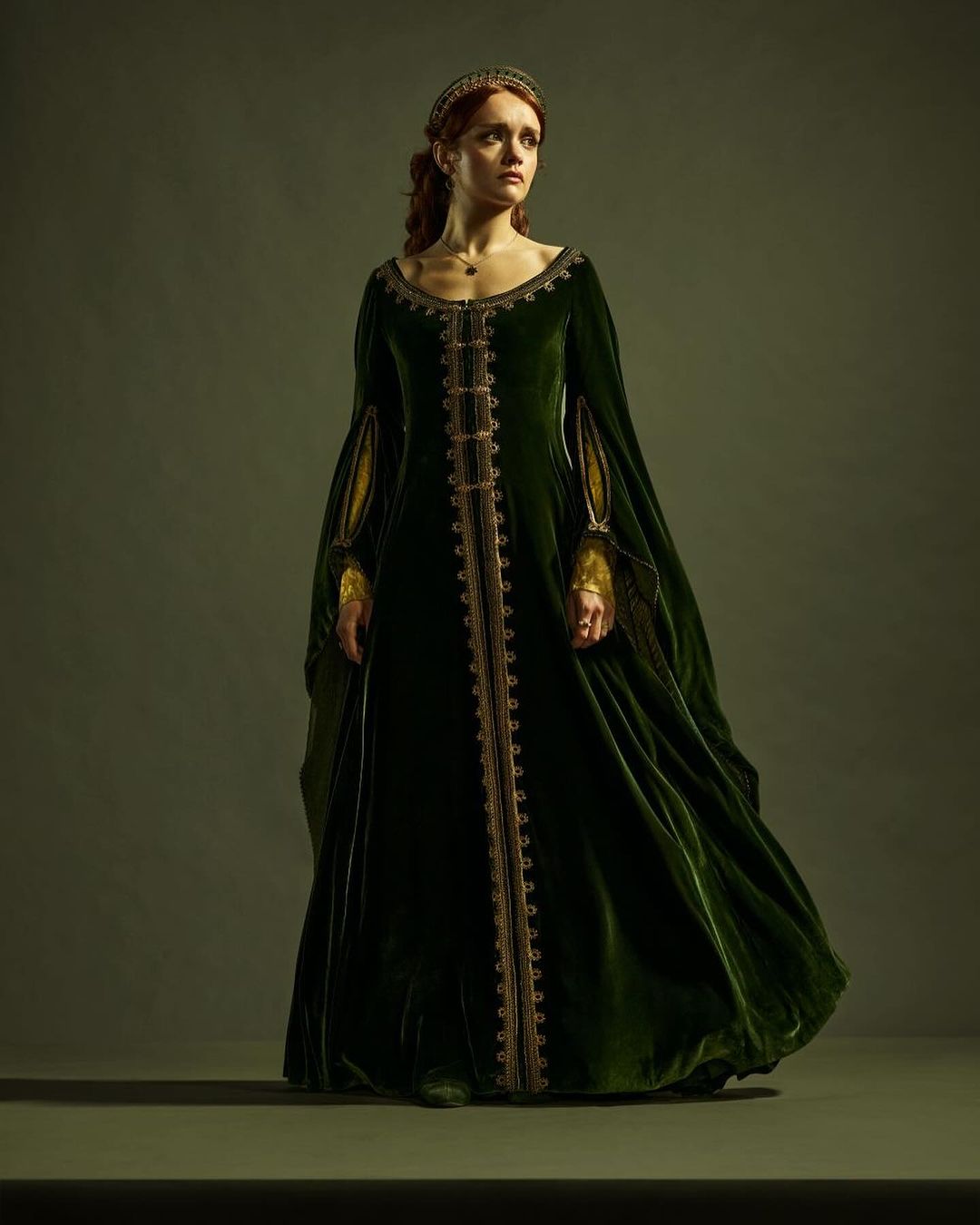 Woman stands in a long, dark green medieval-style gown with gold trim and a jeweled headband, posing in a studio against a solid background.