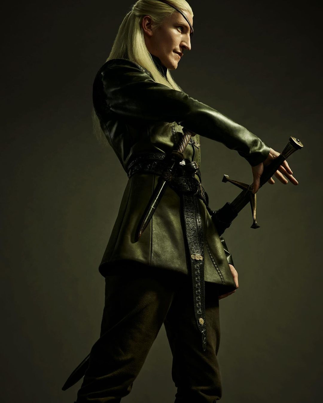 Man with long blond hair and eyepatch poses with sword. He is wearing dark medieval garb in a studio setting with soft lighting.
