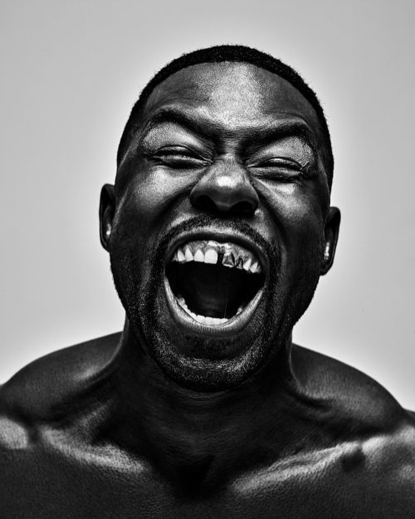 Close-up portrait of an adult Black male with a muscular build and a gold tooth, eyes closed, mouth open in a yell, against a grey background.