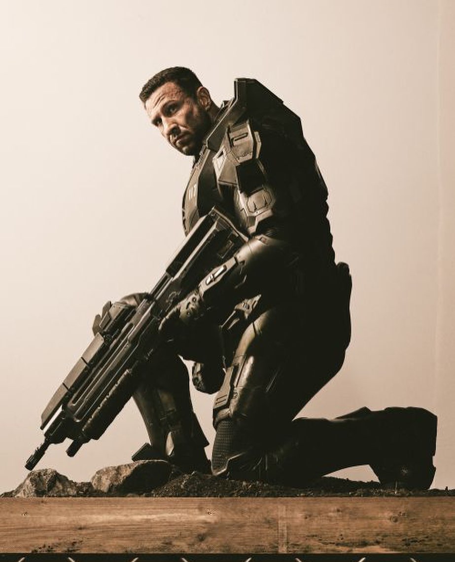 A man in dark futuristic armor kneels holding a large rifle against a light backdrop, creating a striking, intense, and prepared character portrayal.