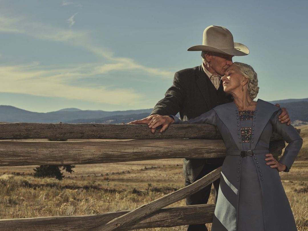 Older adult couple embracing near a wooden fence in a golden field with mountains in the background under a sunny sky.