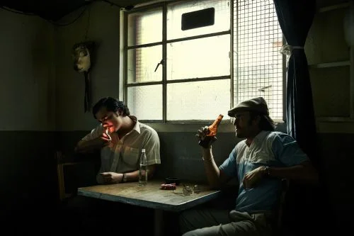 Two men seated at a wooden table in a dimly lit room, one lighting a cigarette while the other holds a beer bottle.
