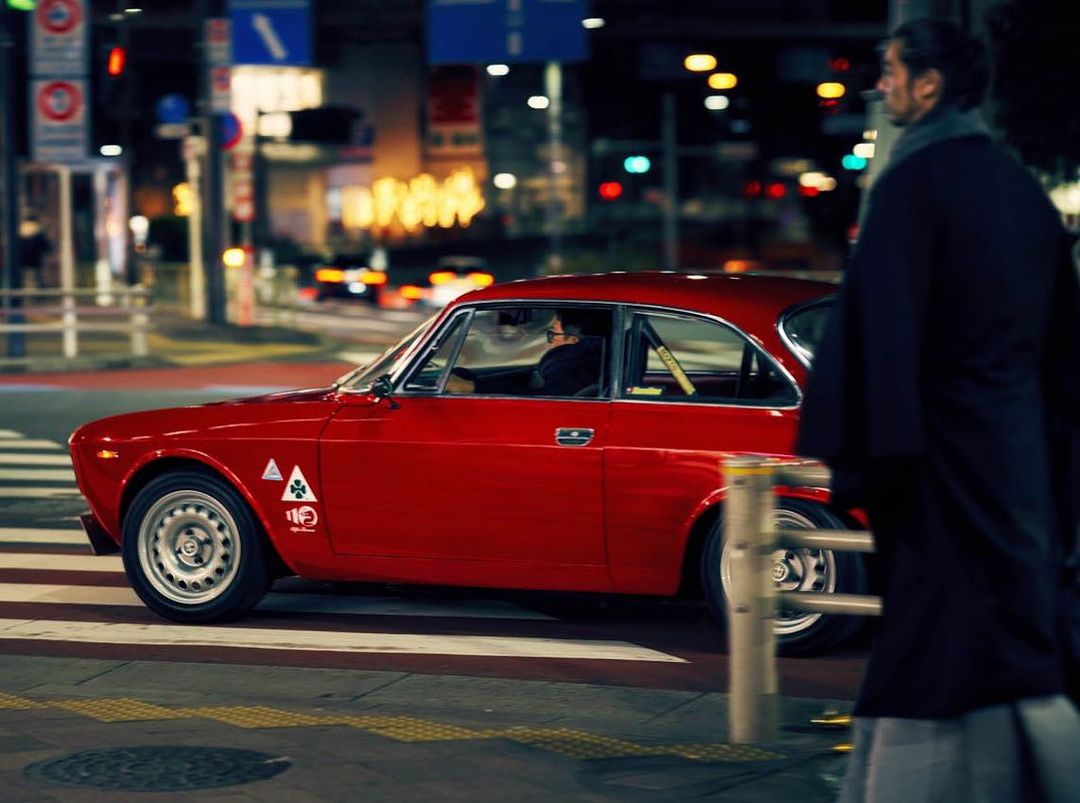 Red Alfa Romeo classic car driving on a city street at night, illuminated by street lights and car headlights with a pedestrian walking nearby.