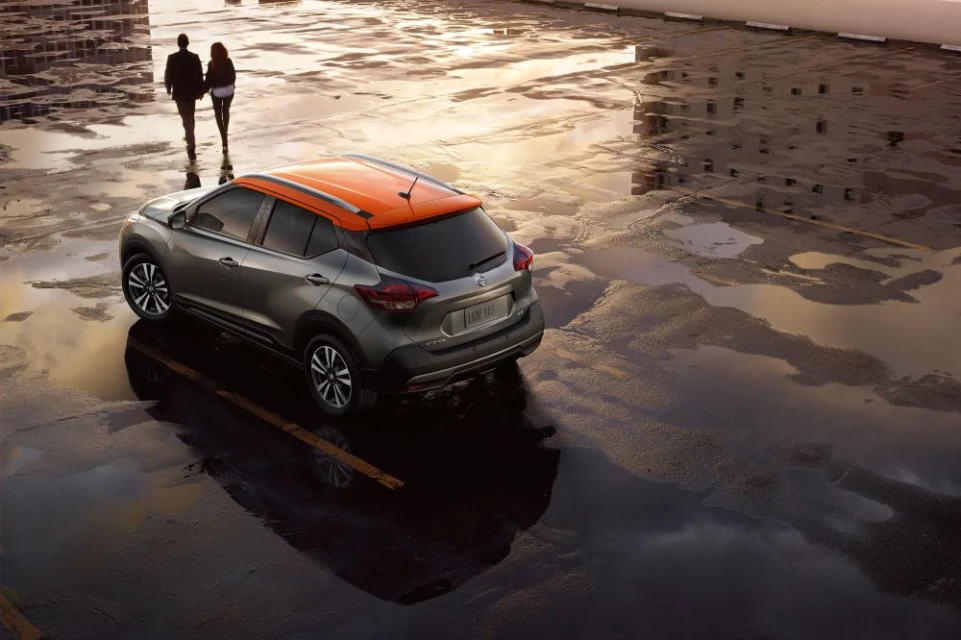 Gray and orange Nissan Kicks crossover on a wet parking lot with reflections, and a couple walking in the background at dusk.