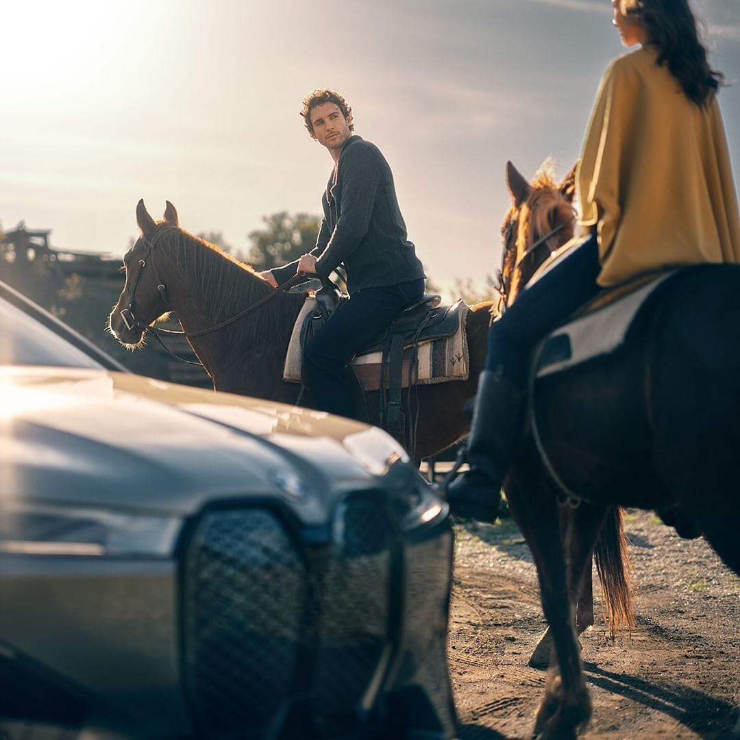 Two individuals on horseback pause near a modern car in a rural setting under warm sunlight, blending equestrian and automotive themes.