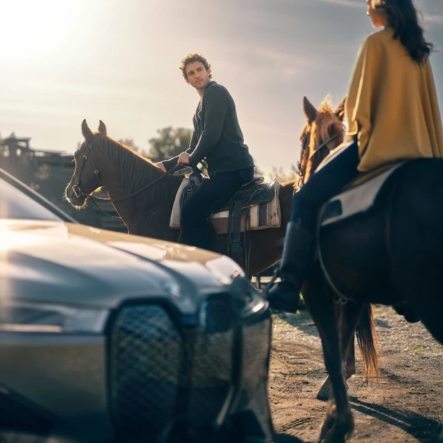 Two individuals on horseback pause near a modern car in a rural setting under warm sunlight, blending equestrian and automotive themes.