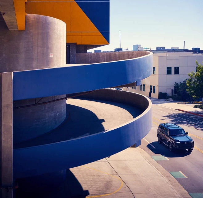 Dark SUV driving on a road that goes around a parking garage with a spiral structure and a building in the background on a sunny day.