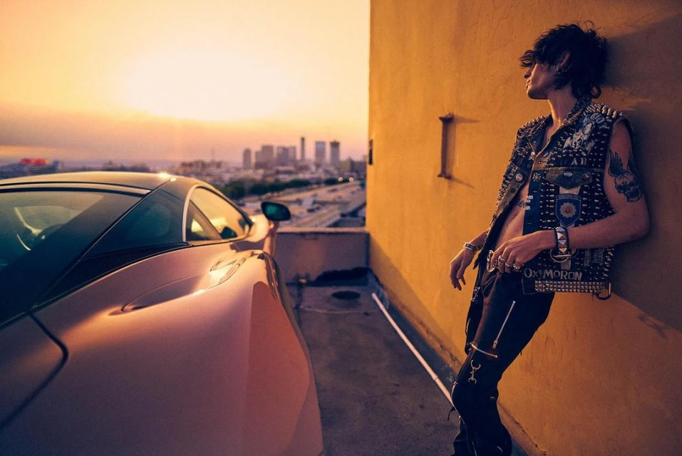 Adult male model poses with a luxury car on a rooftop in an urban setting at sunset, wearing a punk-style studded vest.