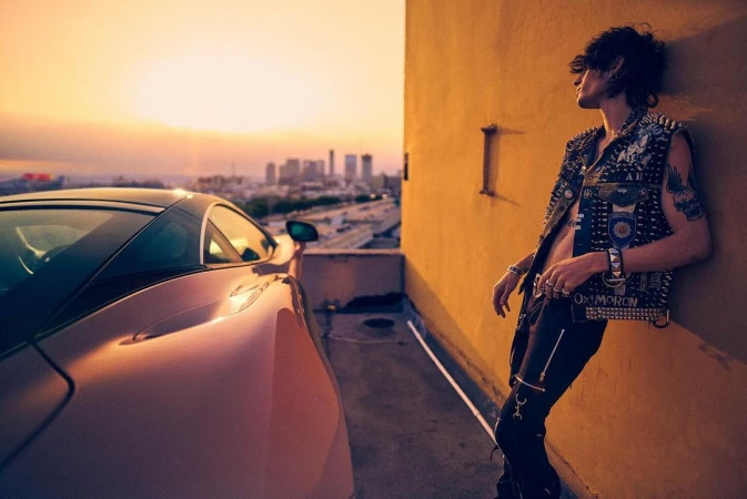 Adult male model poses with a luxury car on a rooftop in an urban setting at sunset, wearing a punk-style studded vest.