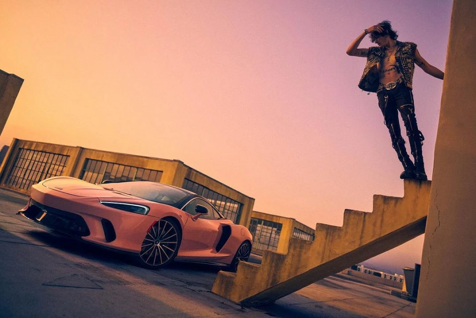 A pink McLaren GT sports car is parked near a man in a studded vest standing on a concrete staircase with urban buildings at sunset.