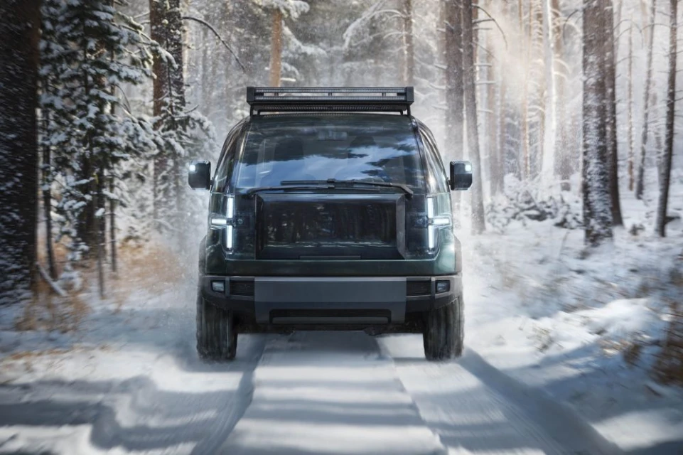 Front view of a dark green van driving on a snowy road through a forest, emphasizing its modern design and winter environment.