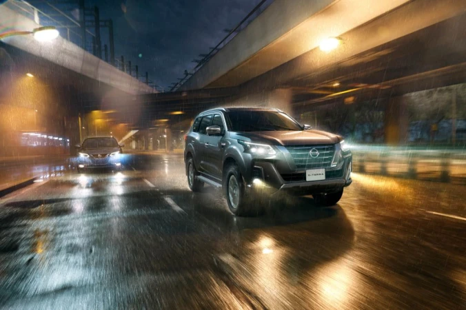 Grey SUV driving on wet asphalt road in the city at night in the rain under overhead bridge with city lights reflecting on the road.