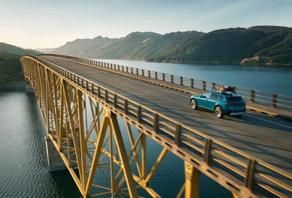 Teal SUV driving on yellow bridge over blue lake with tree-covered mountains in background on sunny day, clear sky, scenic vista.