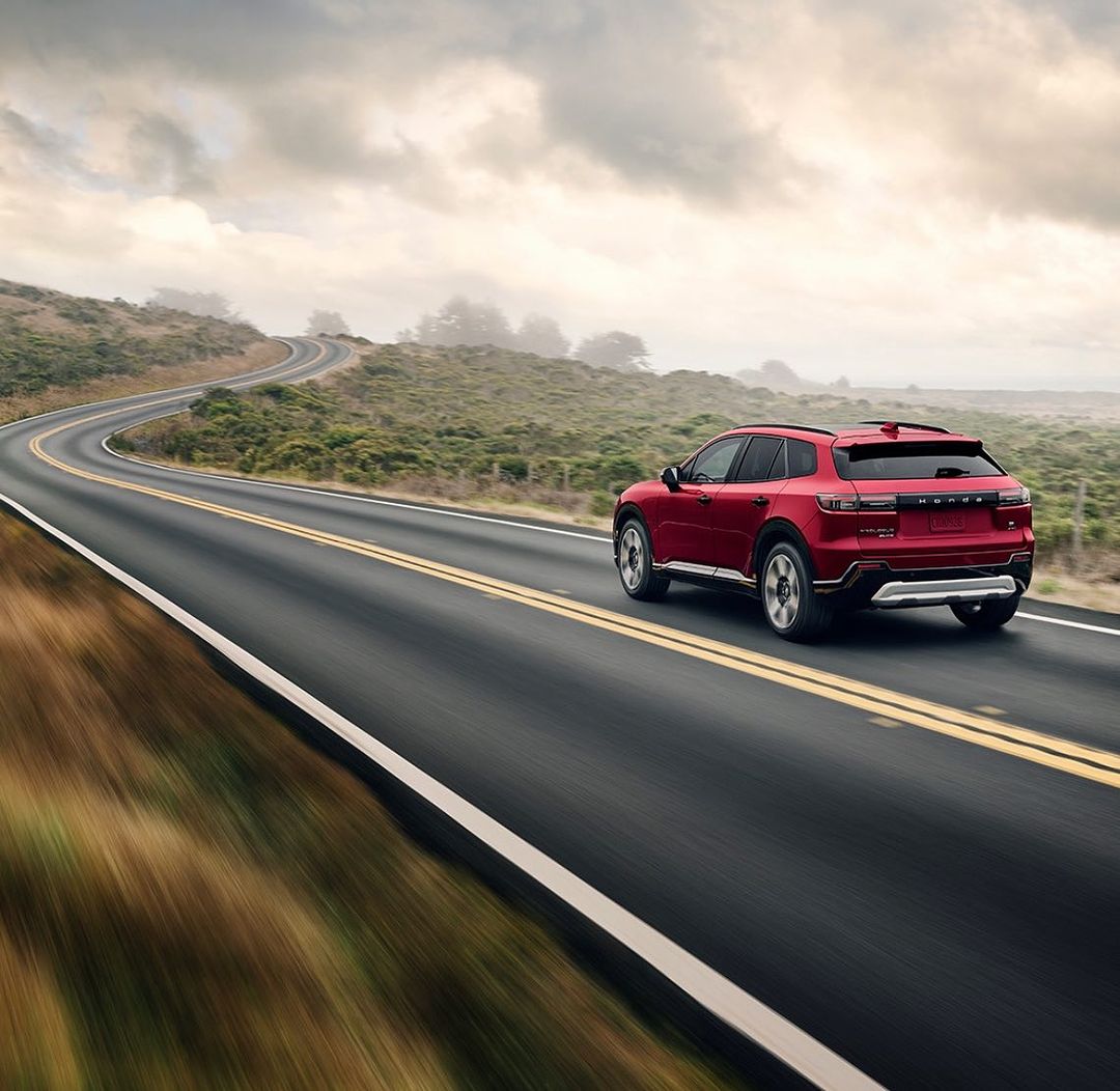 A red SUV drives on a winding coastal road on a cloudy day, with blurred foreground motion adding a sense of speed and adventure.