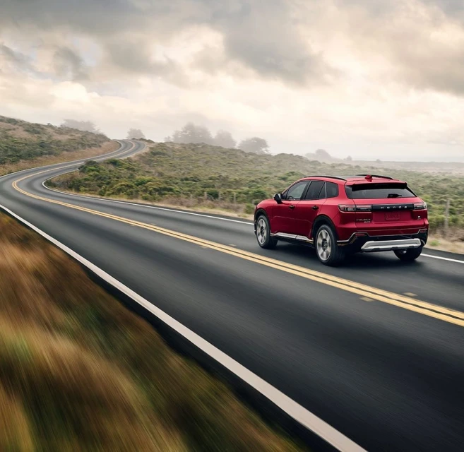 A red SUV drives on a winding coastal road on a cloudy day, with blurred foreground motion adding a sense of speed and adventure.