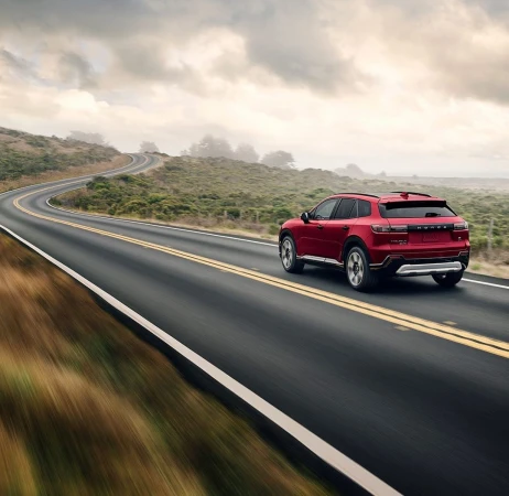 A red SUV drives on a winding coastal road on a cloudy day, with blurred foreground motion adding a sense of speed and adventure.
