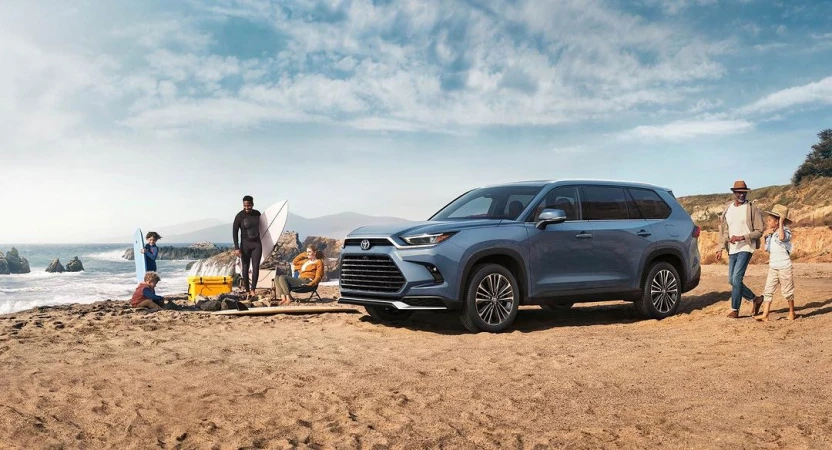 Blue SUV parked on sandy beach with family enjoying seaside; children play, man surfs, and woman relaxes. A sunny, idyllic family moment.