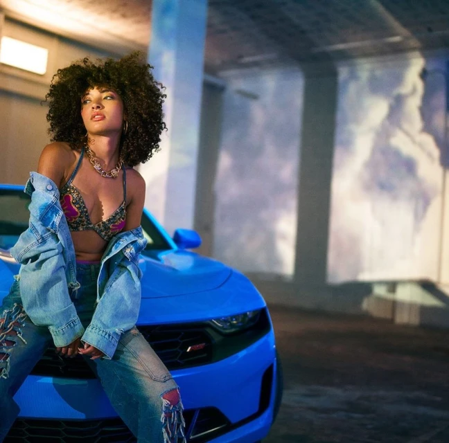 Woman poses on a bright blue Chevrolet Camaro in an indoor setting with soft lighting, wearing a bra top and jeans with a jacket.