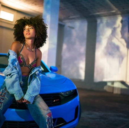 Woman poses on a bright blue Chevrolet Camaro in an indoor setting with soft lighting, wearing a bra top and jeans with a jacket.
