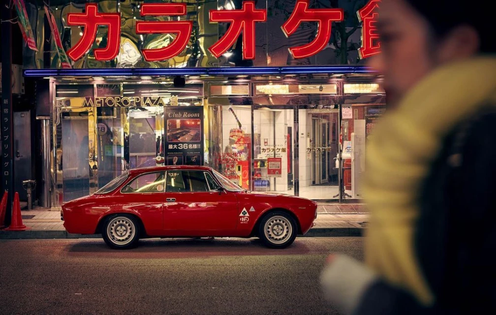 Red Alfa Romeo parked outside a karaoke bar in a Japanese city at night. Neon lights illuminate the street and surrounding buildings.