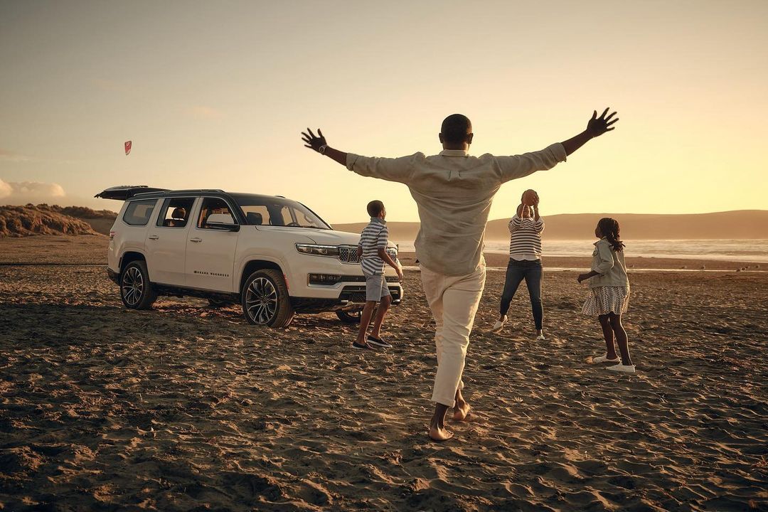 Family enjoys sunset at the beach with a white Jeep Grand Wagoneer. The vehicle is parked near the open ocean and sandy shore.
