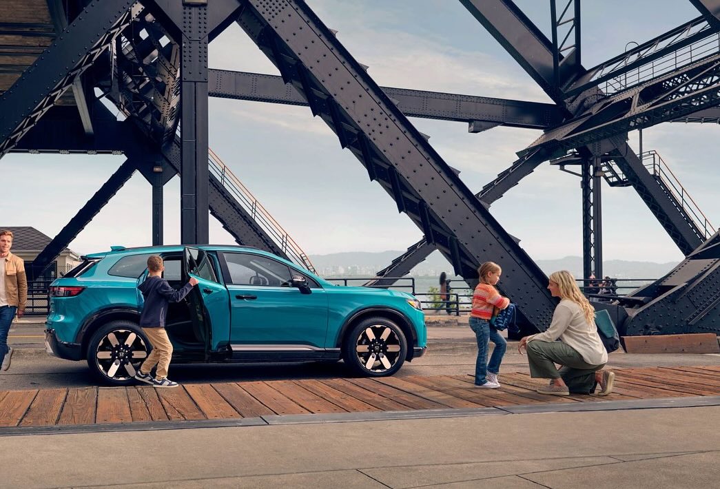 Turquoise electric SUV with family on bridge: child enters car as mother and daughter watch; father walks nearby. Overhead bridge structure.