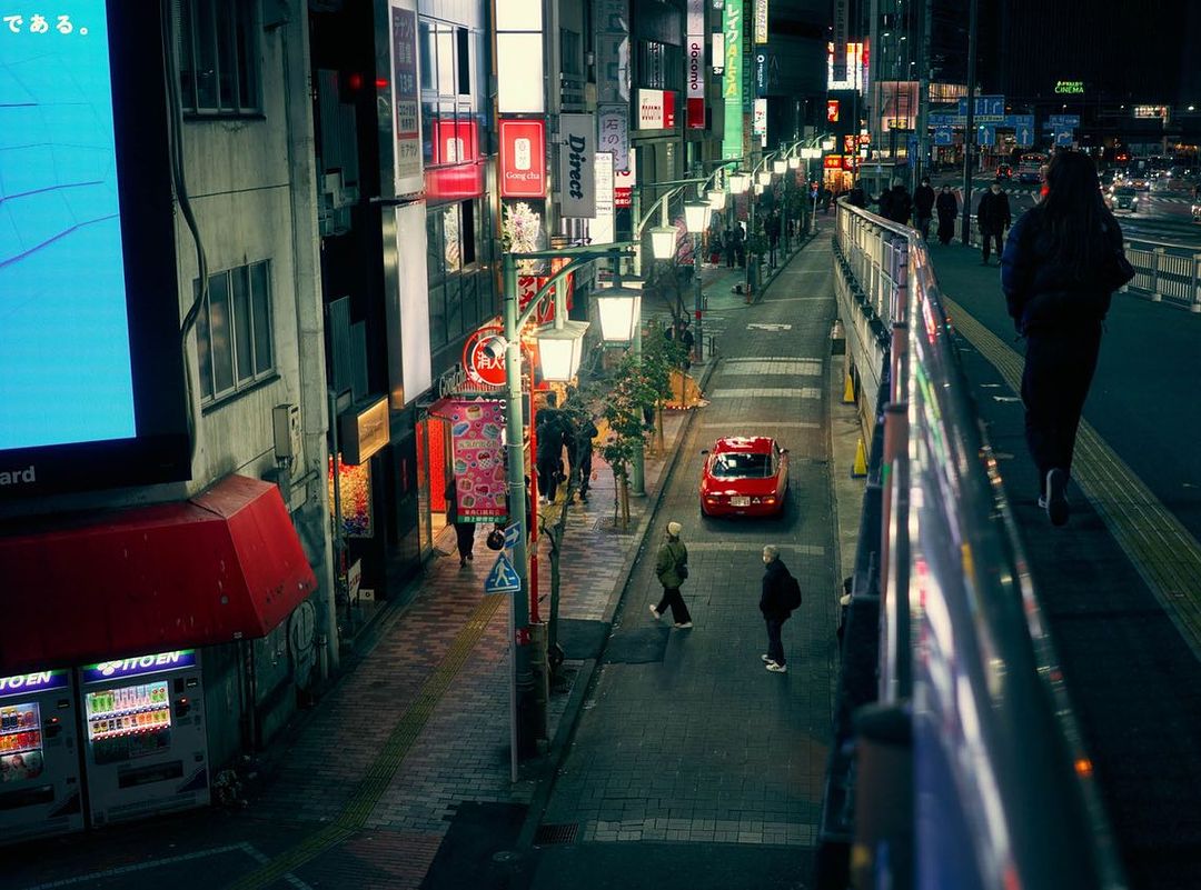 Nighttime urban scene in Japan features pedestrians on a brightly lit street, illuminated buildings, and a red car.