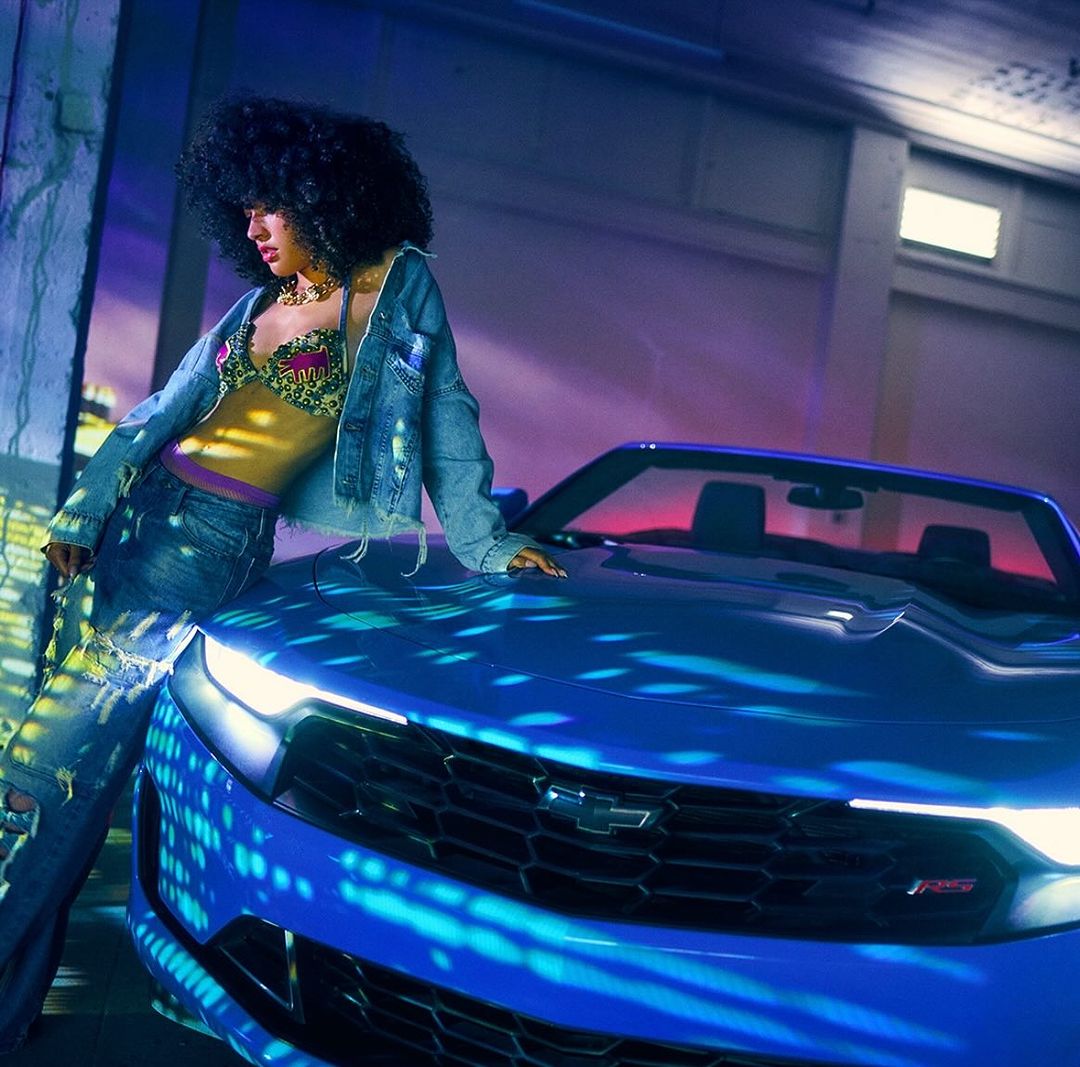 A woman with curly hair in a bra and denim jacket leans on the hood of a blue Chevrolet Camaro convertible in an indoor setting.