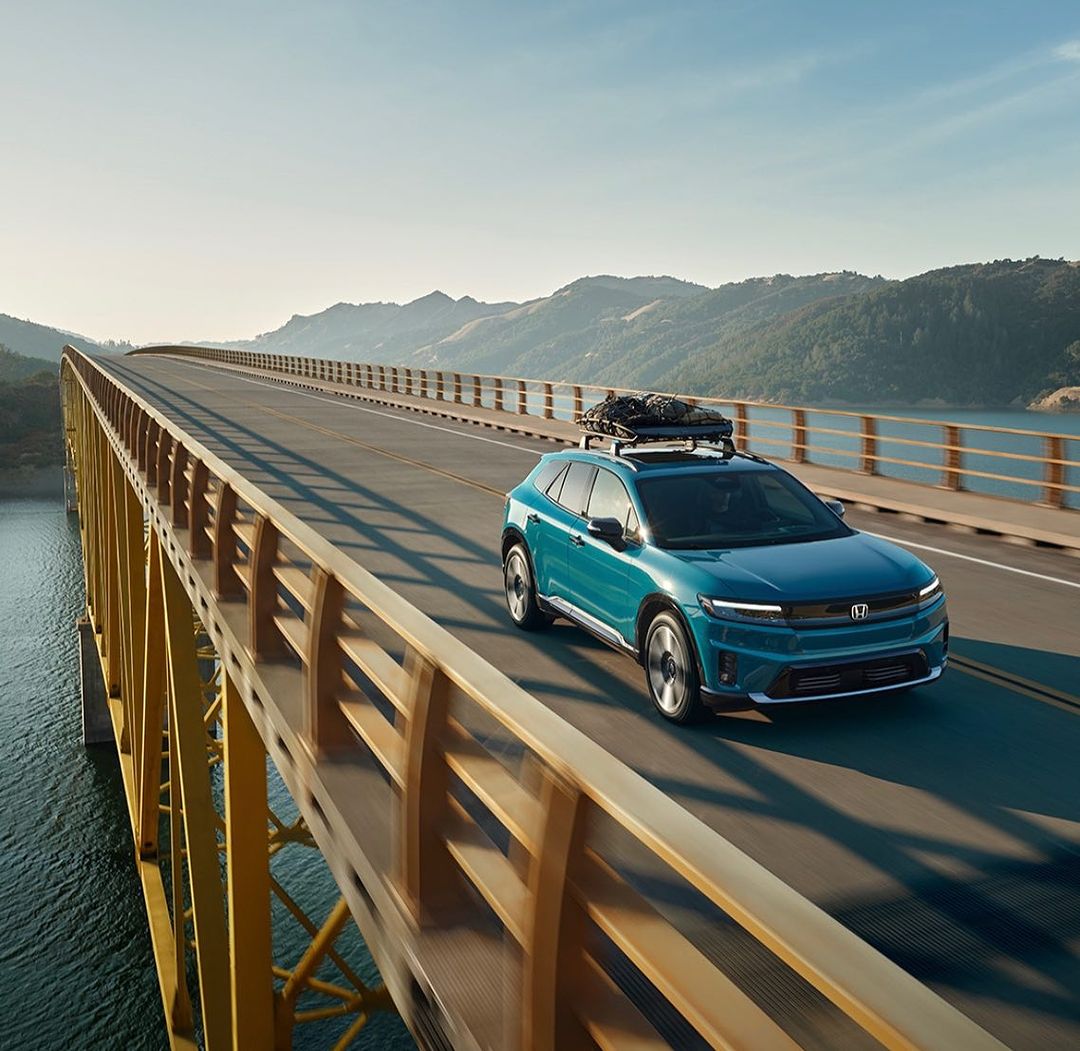 Teal crossover vehicle driving across a yellow steel bridge with mountains and a lake in the background under a clear sky.
