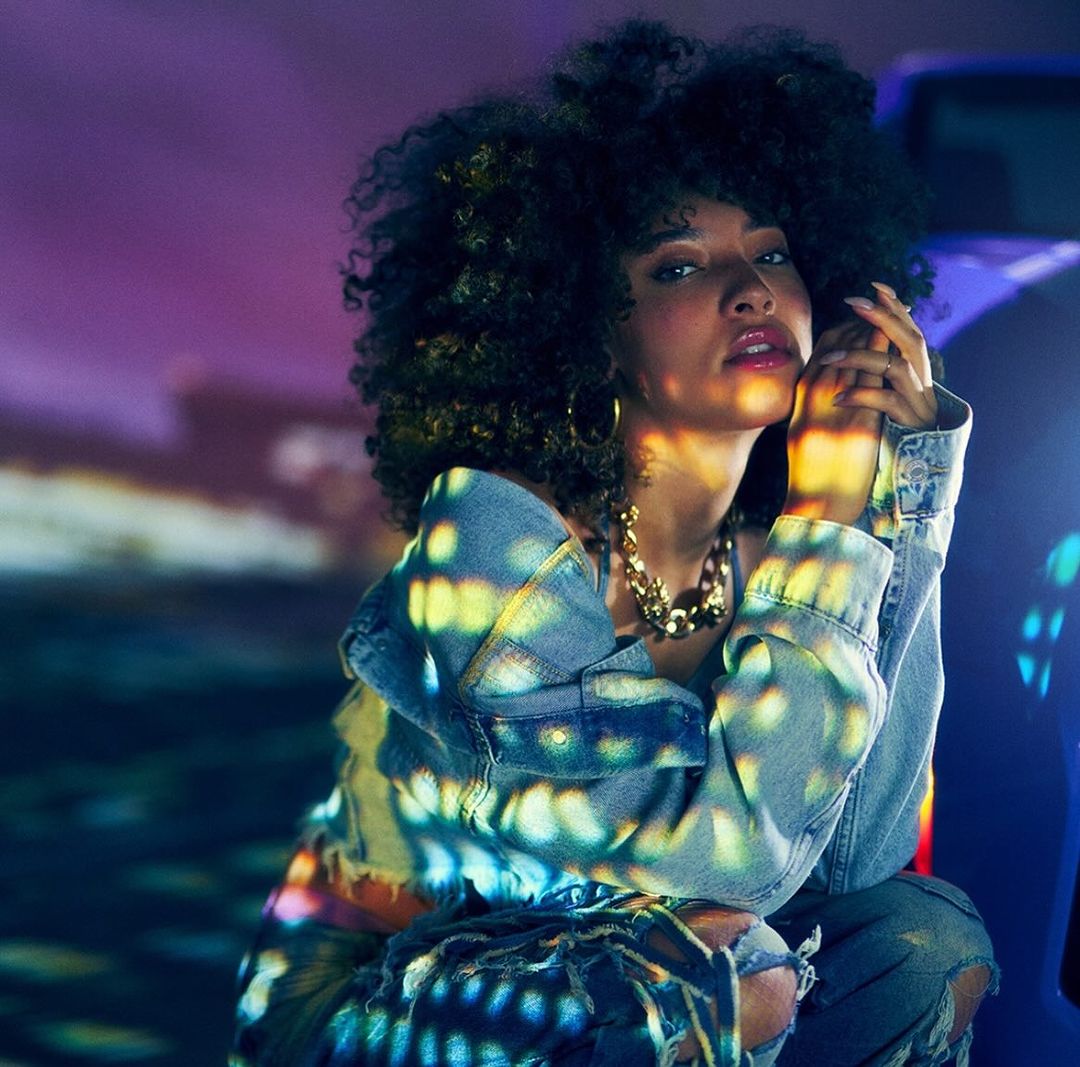 Portrait of a woman with curly hair wearing a denim jacket, gold jewelry, and ripped jeans. She is kneeling in a dimly lit area.