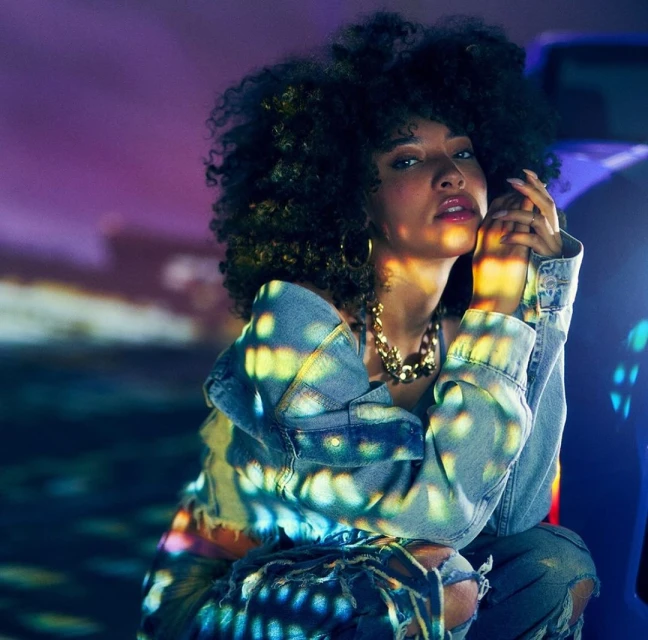 Portrait of a woman with curly hair wearing a denim jacket, gold jewelry, and ripped jeans. She is kneeling in a dimly lit area.