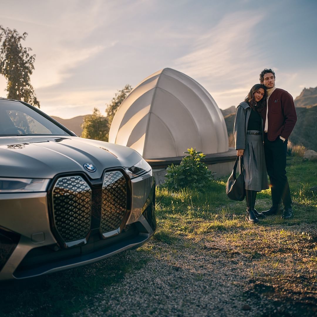 Modern grey BMW SUV parked outdoors with a couple standing nearby and an architectural dome structure in the background, dusk lighting.