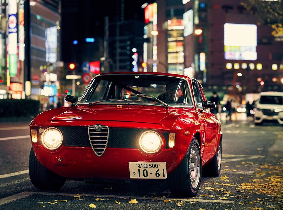 Red Alfa Romeo classic car parked on a city street at night, with illuminated buildings and street lights in the background.