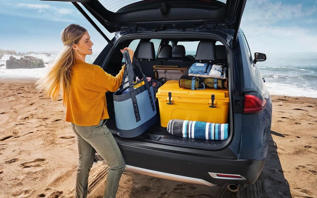 Woman loading a blue tote bag into the trunk of a dark grey SUV parked on a sandy beach with ocean and rock formations in the background.
