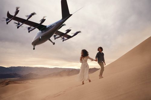 A couple walks hand-in-hand up a sand dune with an electric aircraft above them and distant mountains in the background at dusk.