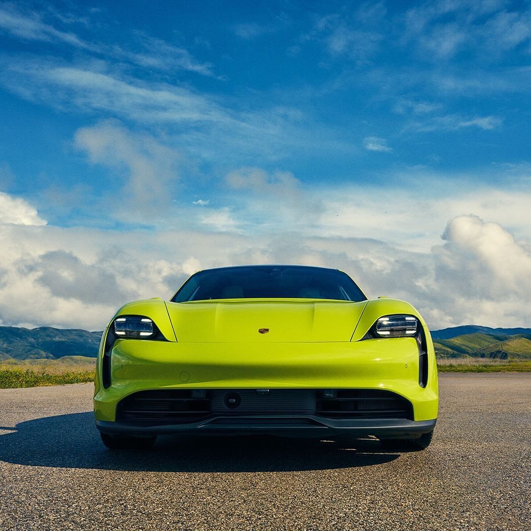 Front view of a vibrant yellow Porsche Taycan parked on an empty asphalt road with a backdrop of a blue sky and distant hills.