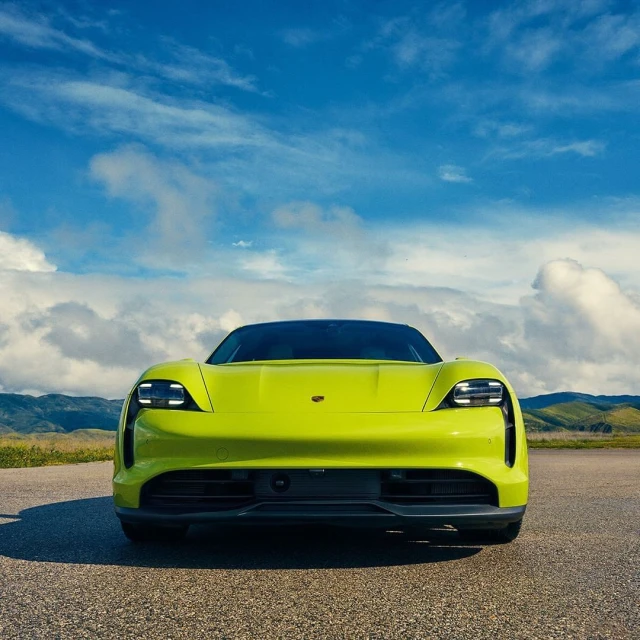Front view of a vibrant yellow Porsche Taycan parked on an empty asphalt road with a backdrop of a blue sky and distant hills.