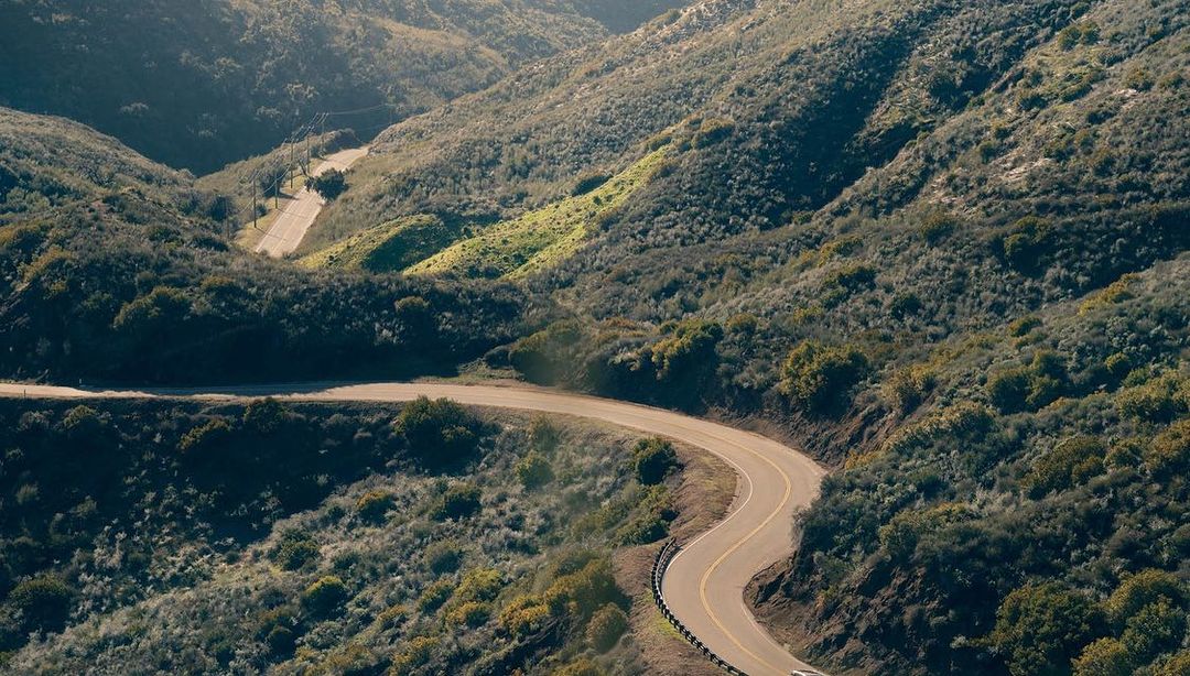 High-angle shot showcases a winding road with yellow lines curving through a hilly, natural landscape covered with green vegetation.