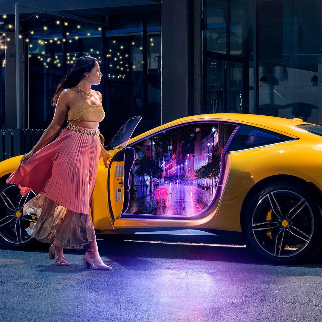 A fashionable woman in a pleated skirt standing next to a yellow sports car with its door open, reflecting an urban city street.