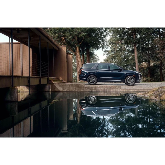Dark blue SUV parked near a modern house with a reflective pond showing the car's image, surrounded by trees and gravel.