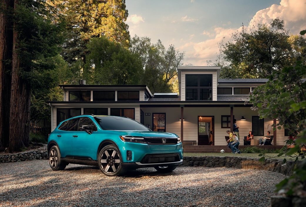 A light-blue Honda Prologue electric SUV is parked in front of a modern two-story home with children and adults on the porch.