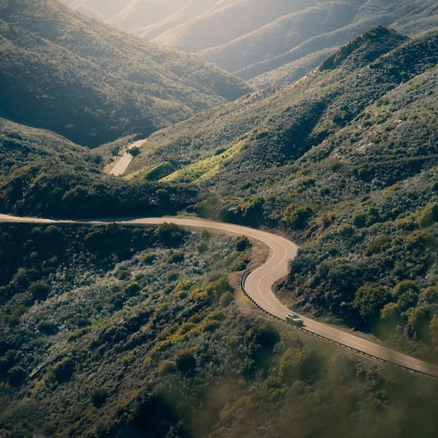 Aerial view of a car driving on a winding road through lush, green hills under soft sunlight. Scenic landscape with a sense of adventure.