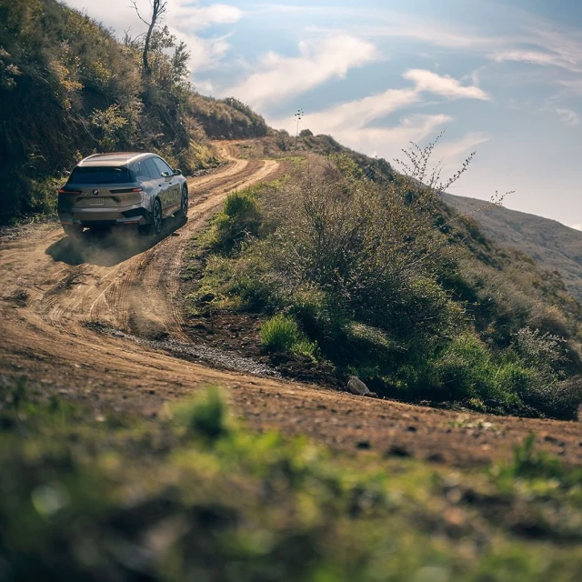 SUV driving on a dirt road with hills in the background on a sunny day. The vehicle is kicking up dust as it navigates the path.